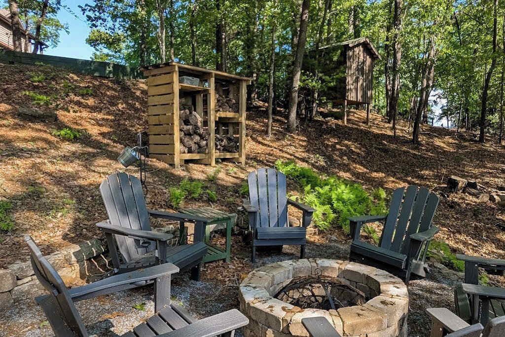 A forested area with a fire pit, Adirondack chairs, and a wooden structure.