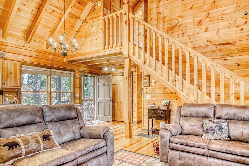 A wooden cabin interior with a staircase, leather sofas, and a chandelier.