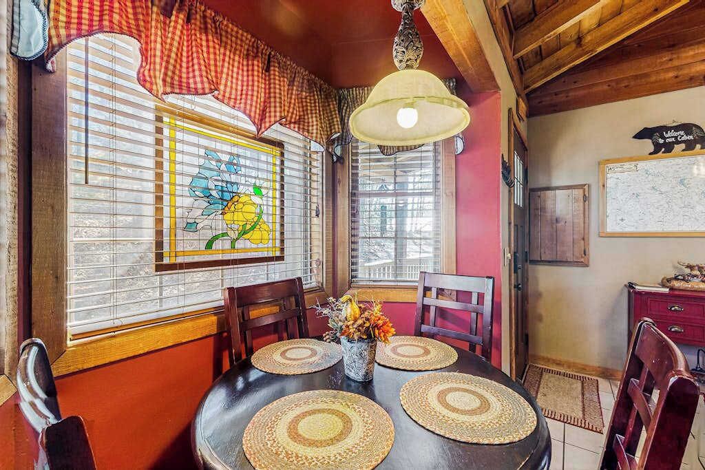 A dining area with a wooden table, four chairs, a vase with flowers, and a stained glass window.