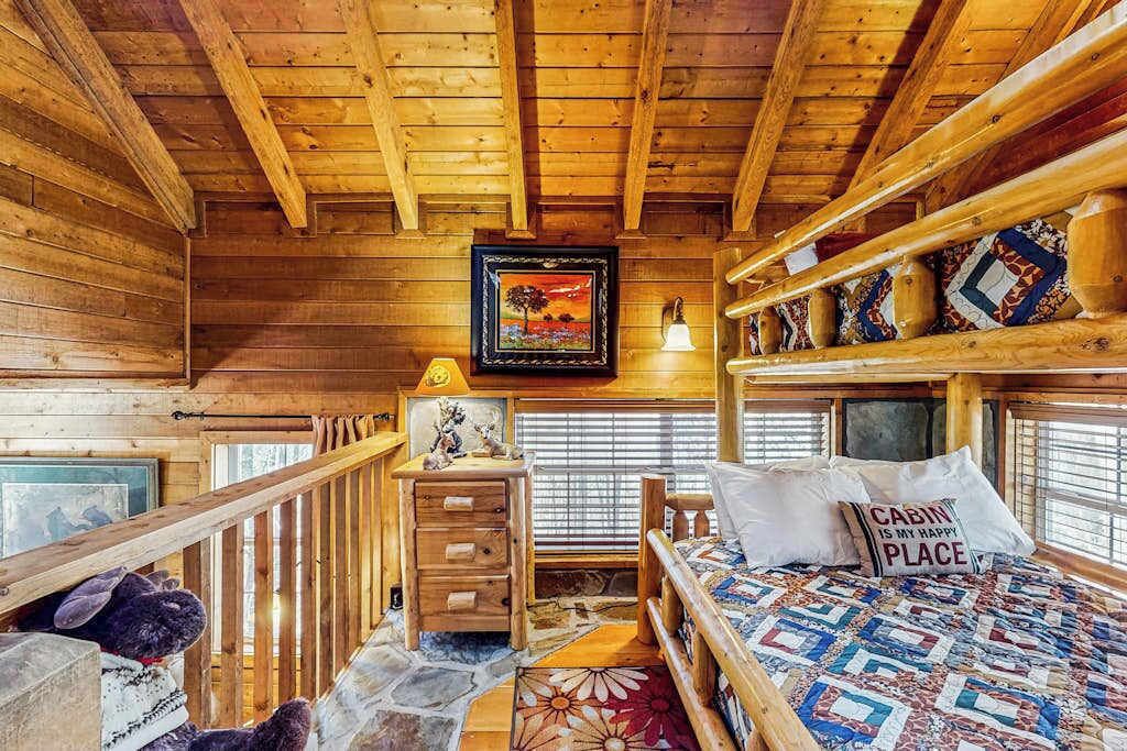A cabin interior with a wooden ceiling, a bed with a quilt, a dresser, a framed picture, and a window with blinds.