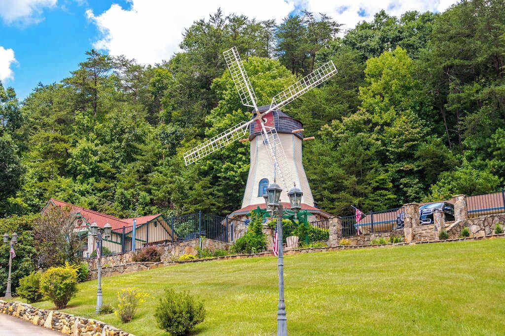 A windmill with a stone base, surrounded by a stone wall, and a small building with a red roof in the foreground.