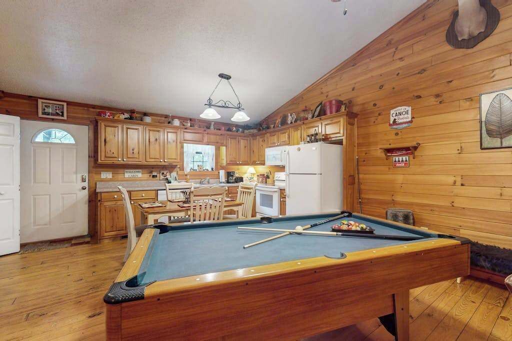 Una mesa de billar en una habitación con paredes de madera y una zona de cocina al fondo.