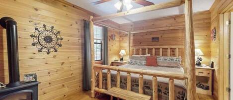A log cabin bedroom with a bed, a ceiling fan, a fireplace, and a window.