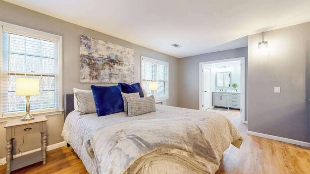 Une chambre à coucher avec un grand lit, deux tables de chevet, une commode, une fenêtre avec des stores, un tableau au mur et une salle de bain visible à travers une porte ouverte.