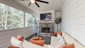 Una habitación con chimenea de piedra, ventilador de techo, televisor sobre la chimenea y un ventanal grande.