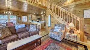 A cabin interior with a wooden staircase, a sofa, a patterned armchair, a small table with a lamp, and a kitchen area. - Dogwood Overlook Cabin (Mineral Bluff)
