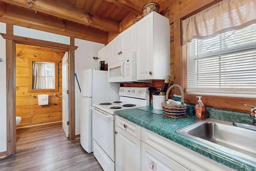 Ein Interieur einer Holzhütte mit Küchenzeile, die weiße Geräte, ein Spülbecken und ein Fenster mit Jalousien zeigt.