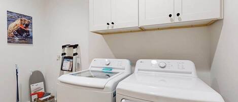 A laundry room with two white front-loading washing machines, a small table with a red object, a mop, and a wall-mounted laundry scale.