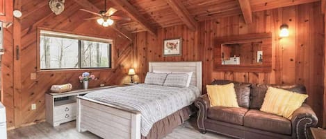 A wooden cabin bedroom with a bed, a wooden dresser, a brown leather sofa, a ceiling fan, and a window.