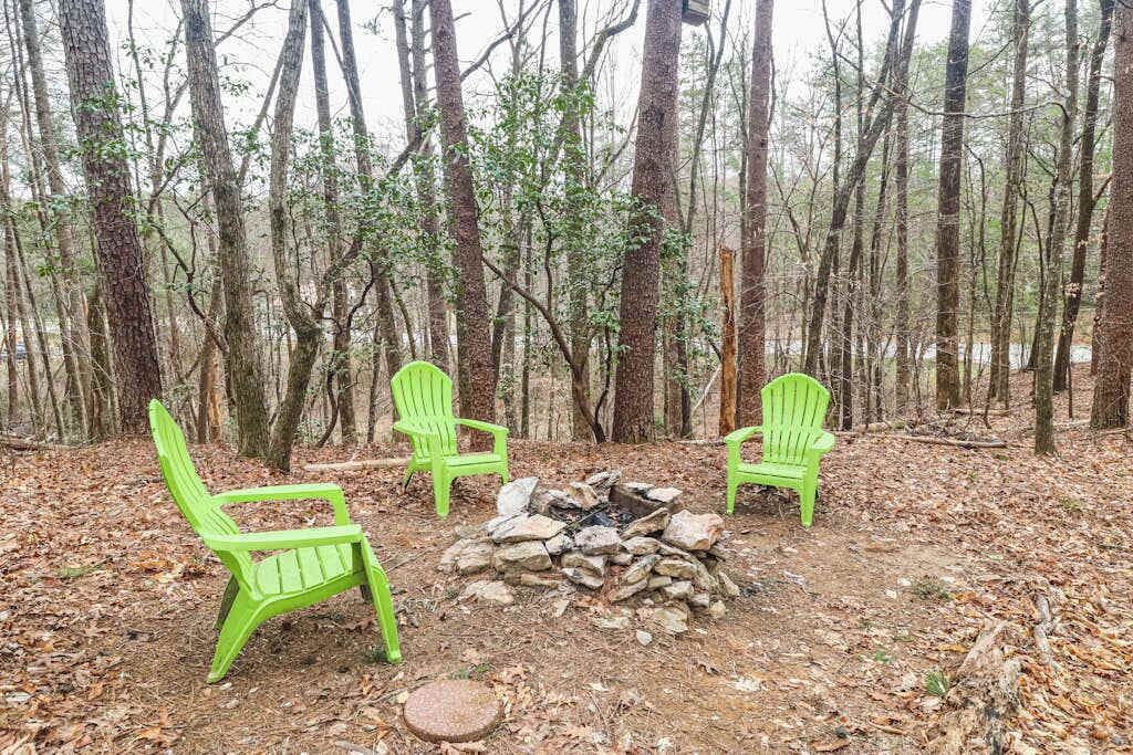 Three green Adirondack chairs arranged around a stone fire pit in a forested area.
