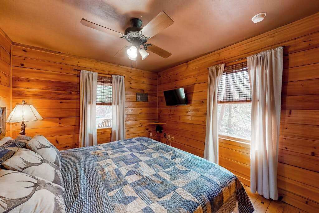 A wooden cabin room with a bed, ceiling fan, TV, and windows with curtains.