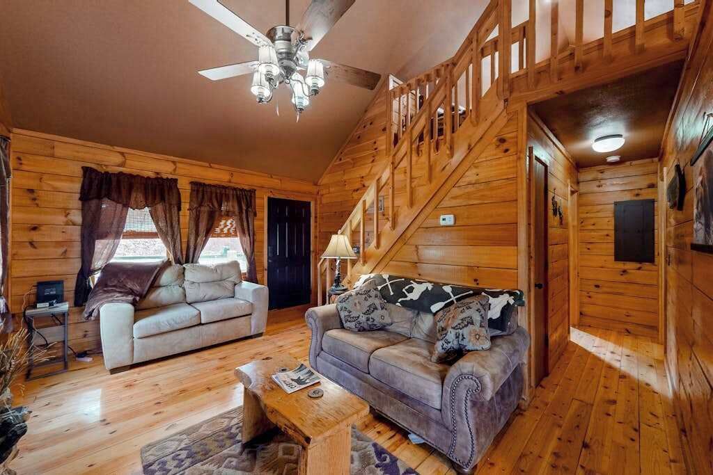 A wooden cabin interior with two sofas, a ceiling fan, and a staircase.