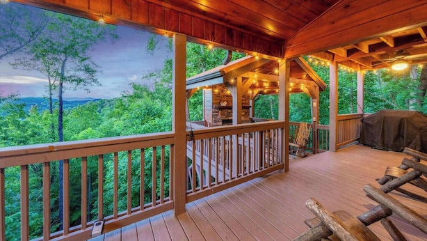 A wooden deck with a railing, a covered seating area, and a view of a forested landscape. - Red Oak Luxe Mountain Cabin (Blue Ridge)