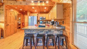 A wooden cabin interior with a kitchen area, bar stools, and a stone island. - Red Oak Luxe Mountain Cabin (Blue Ridge)