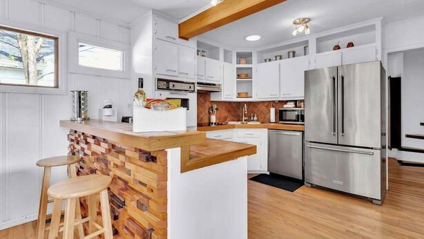 A modern kitchen with white cabinetry, a wooden island with stools, a stainless steel refrigerator, and a wooden ceiling beam. - EnDeering Retreat Stay (Atlanta)