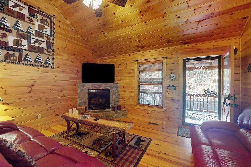 A log cabin interior with a stone fireplace, wooden walls, a leather sofa, and a television.