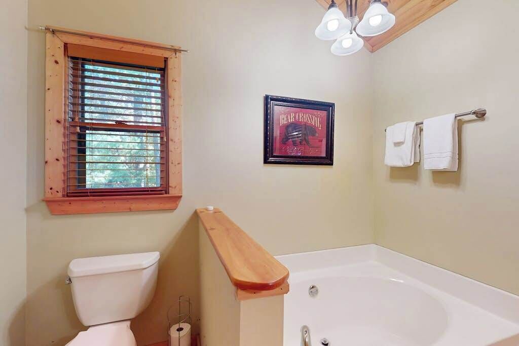 Une salle de bain comprenant des toilettes, une baignoire, une fenêtre avec des stores, un tableau encadré au mur et un porte-serviettes.