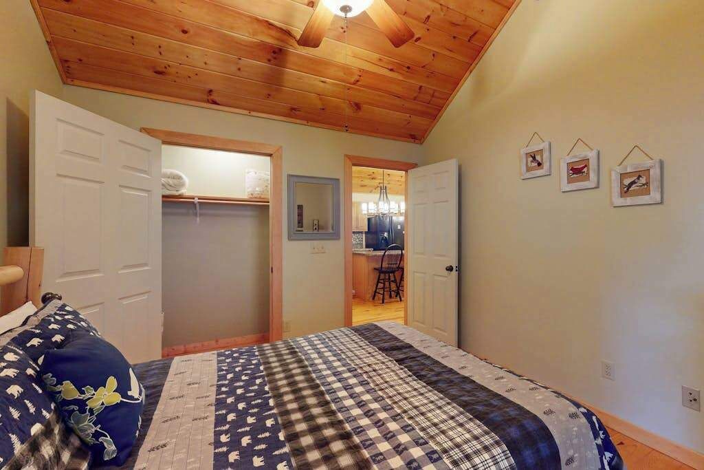 A bedroom with a wooden ceiling, a bed with a patterned bedspread, a closet, and a door leading to a kitchen area.