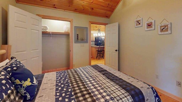 Un dormitorio con techo de madera, una cama con colcha estampada, un armario y una puerta que da a una zona de cocina.