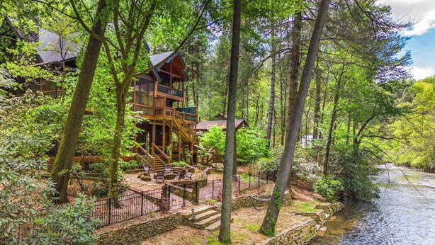 Une cabane dotée d’une terrasse et d’un espace extérieur aménagé, située au bord d’une rivière et entourée d’arbres.