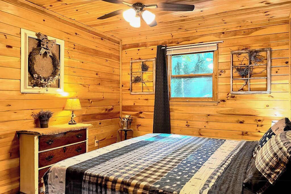 A wooden cabin bedroom with a bed, dresser, lamp, and ceiling fan.
