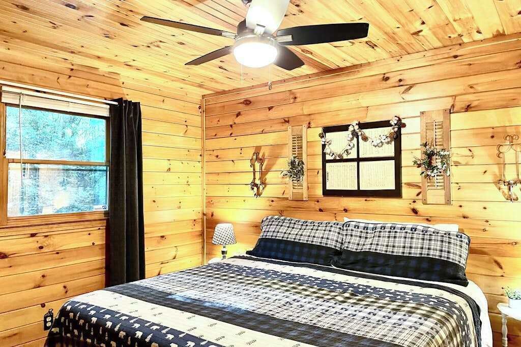 Un dormitorio de cabaña de madera con una cama, un ventilador de techo, una ventana con cortinas y adornos en la pared.