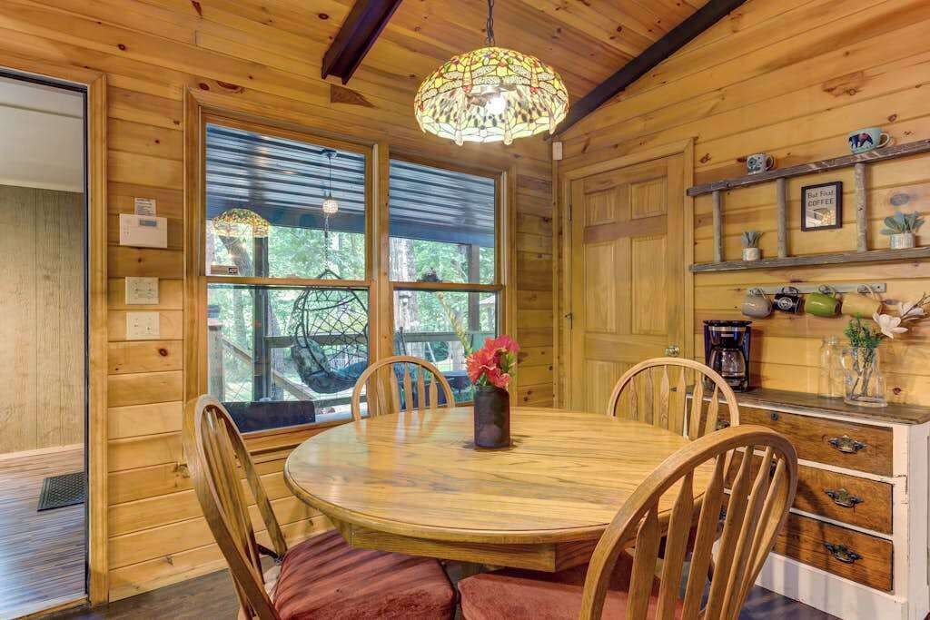 A wooden cabin interior with a round dining table, chairs, a chandelier, and a window with a view of trees.