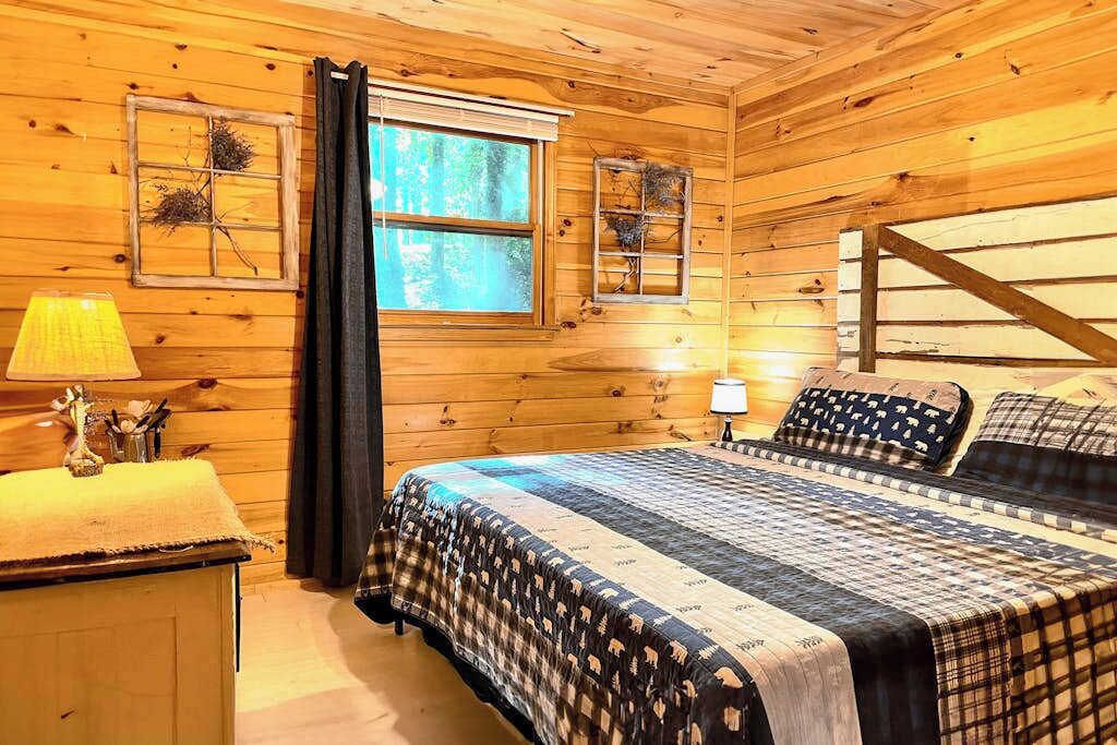A wooden cabin room with a bed, two windows, a lamp, and a small table.