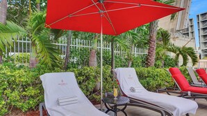 Zona de piscina con sillones de descanso, una sombrilla roja y una pequeña mesa con una bebida.
