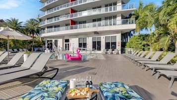 Un área junto a la piscina con sillones, una mesa con comida y bebidas, y un edificio con balcones.
