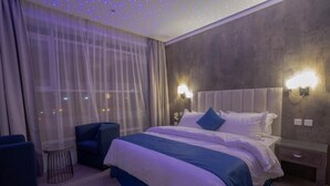 A hotel room with a large bed, two bedside tables, a chair, a window with sheer curtains, and a ceiling with small lights.