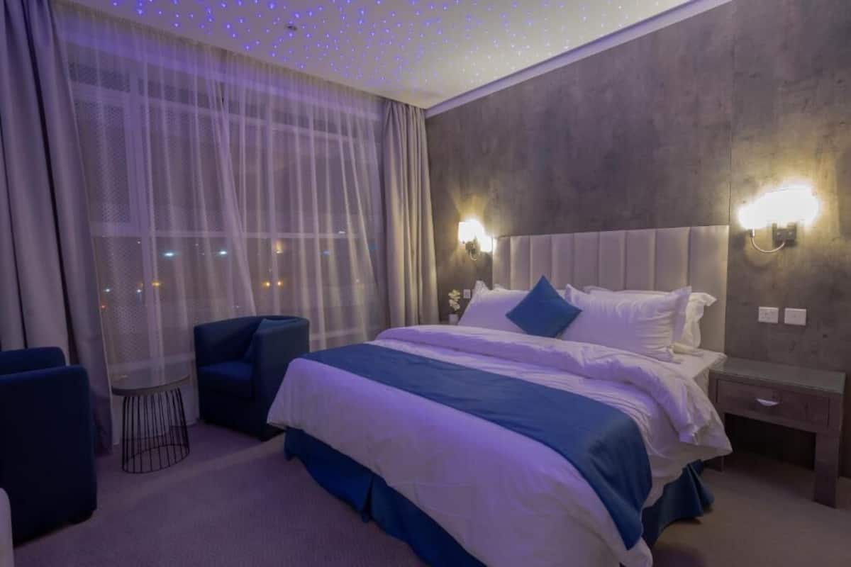 A hotel room with a large bed, two bedside tables, a chair, a window with sheer curtains, and a ceiling with small lights.