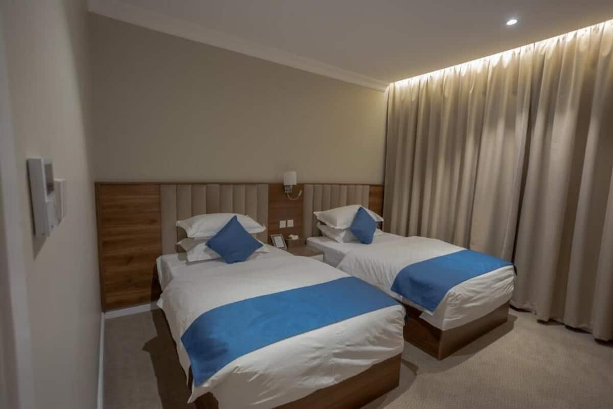 A hotel room with two beds, a wooden headboard, a nightstand, a lamp, and a large window with curtains.