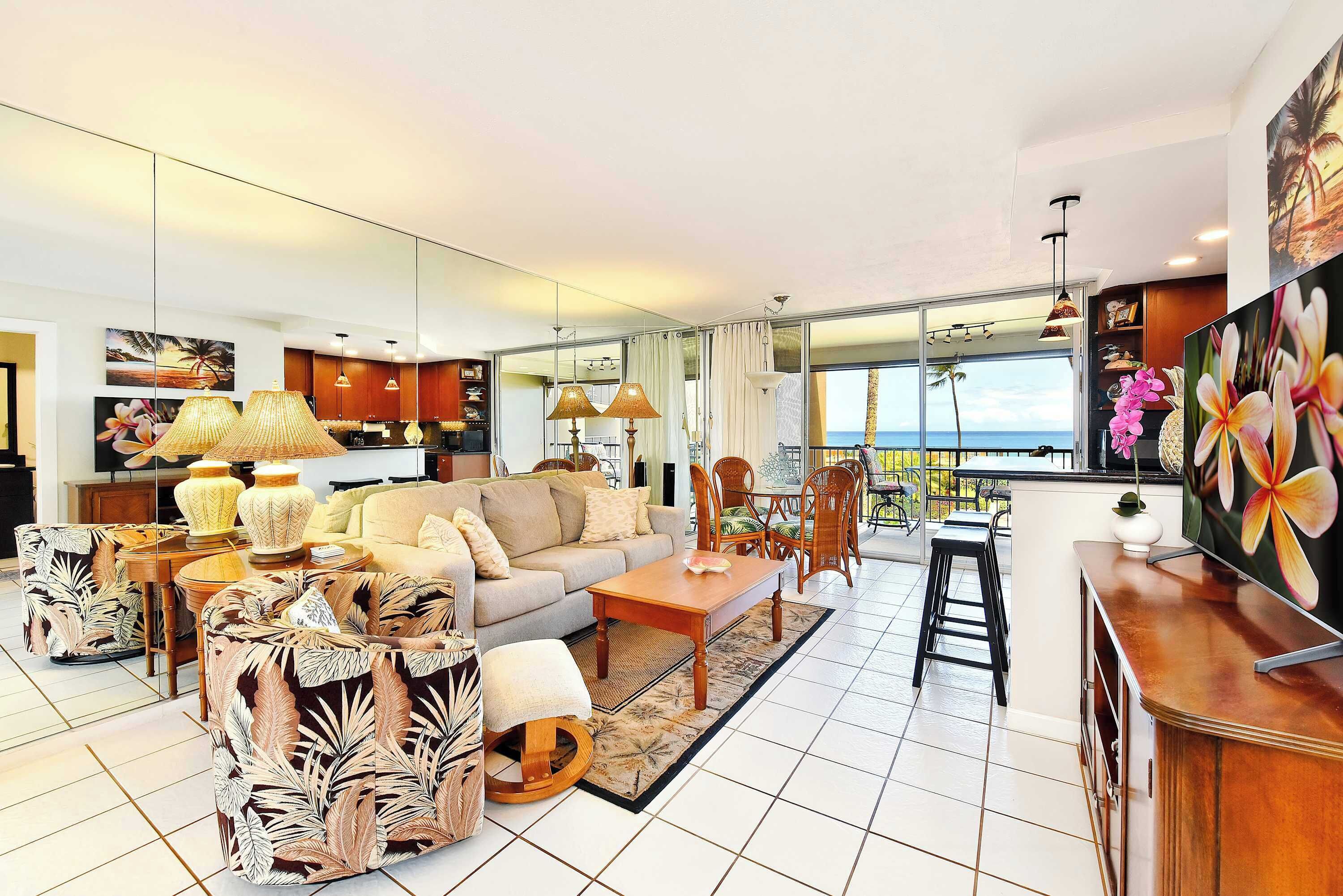 A spacious living area with a sofa, coffee table, and chairs, a kitchen in the background, and a large window with a view of the sea.