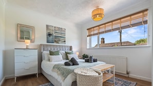 Ein Schlafzimmer mit Bett, Nachttisch, Lampe, Fenster mit Jalousien und Blick auf ein Gebäude.