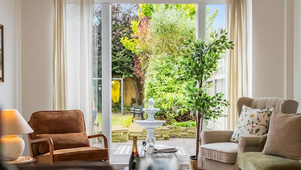 Ein Zimmer mit einer Couch, einem Sessel, einem Tisch mit einer Flasche und Blick auf einen Garten mit einem Springbrunnen.