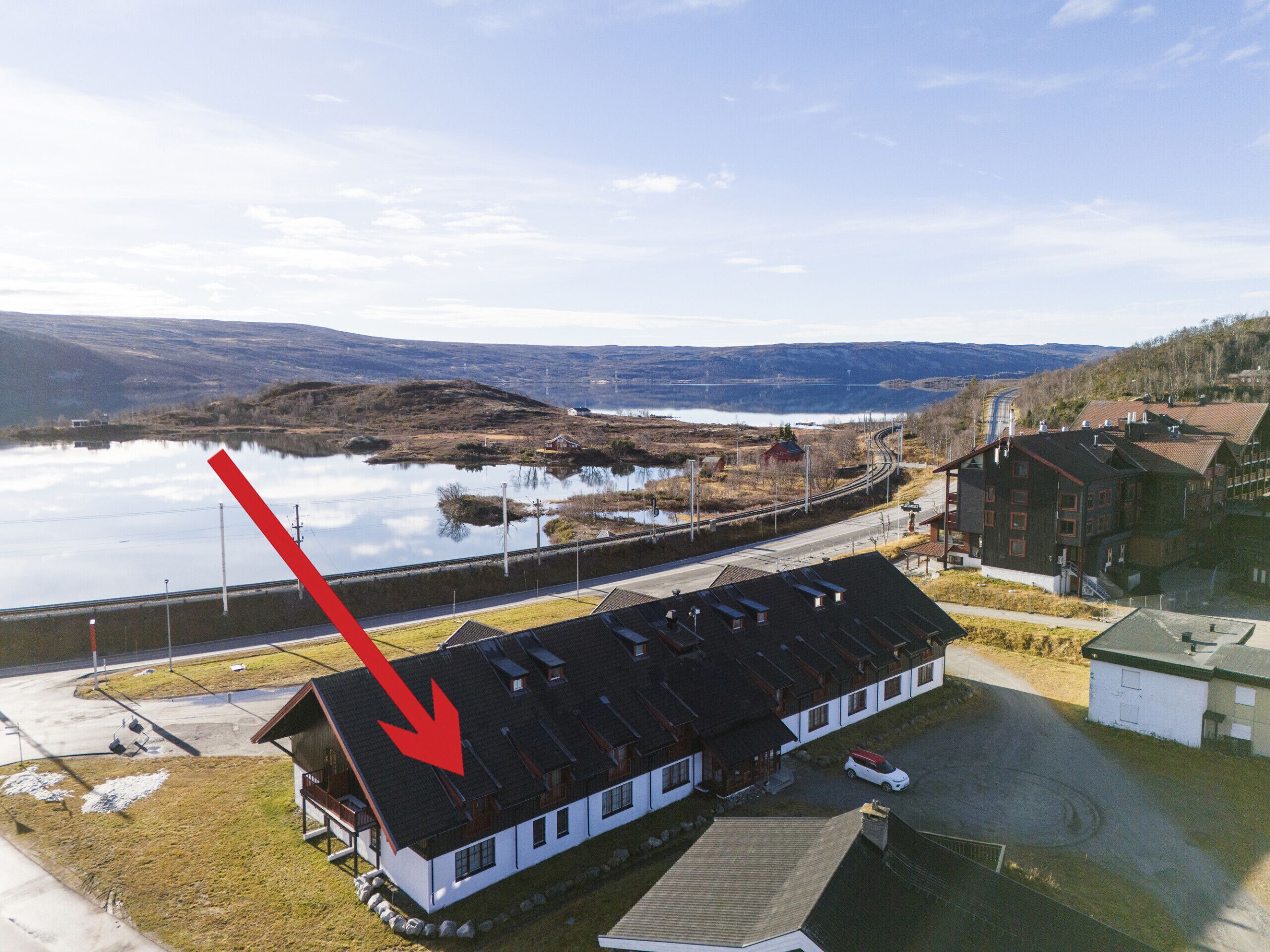 A building with a sloped roof, a parking area, and a view of a lake and mountains in the background.