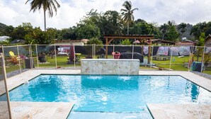 A rectangular swimming pool with a stone border, surrounded by a fence, and a wooden pergola in the background.