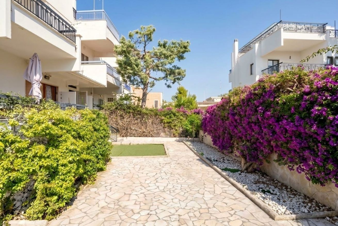 Zona residenziale con vialetto lastricato, edifici bianchi e piante fiorite nei toni del verde e del viola.