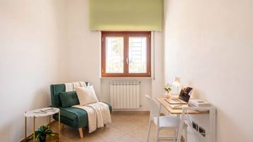 Ein kleiner, gut beleuchteter Raum mit einem grünen Sofa, einem kleinen runden Tisch mit einer Pflanze, einem weißen Schreibtisch mit einem Laptop und einem Heizkörper unter einem Fenster.