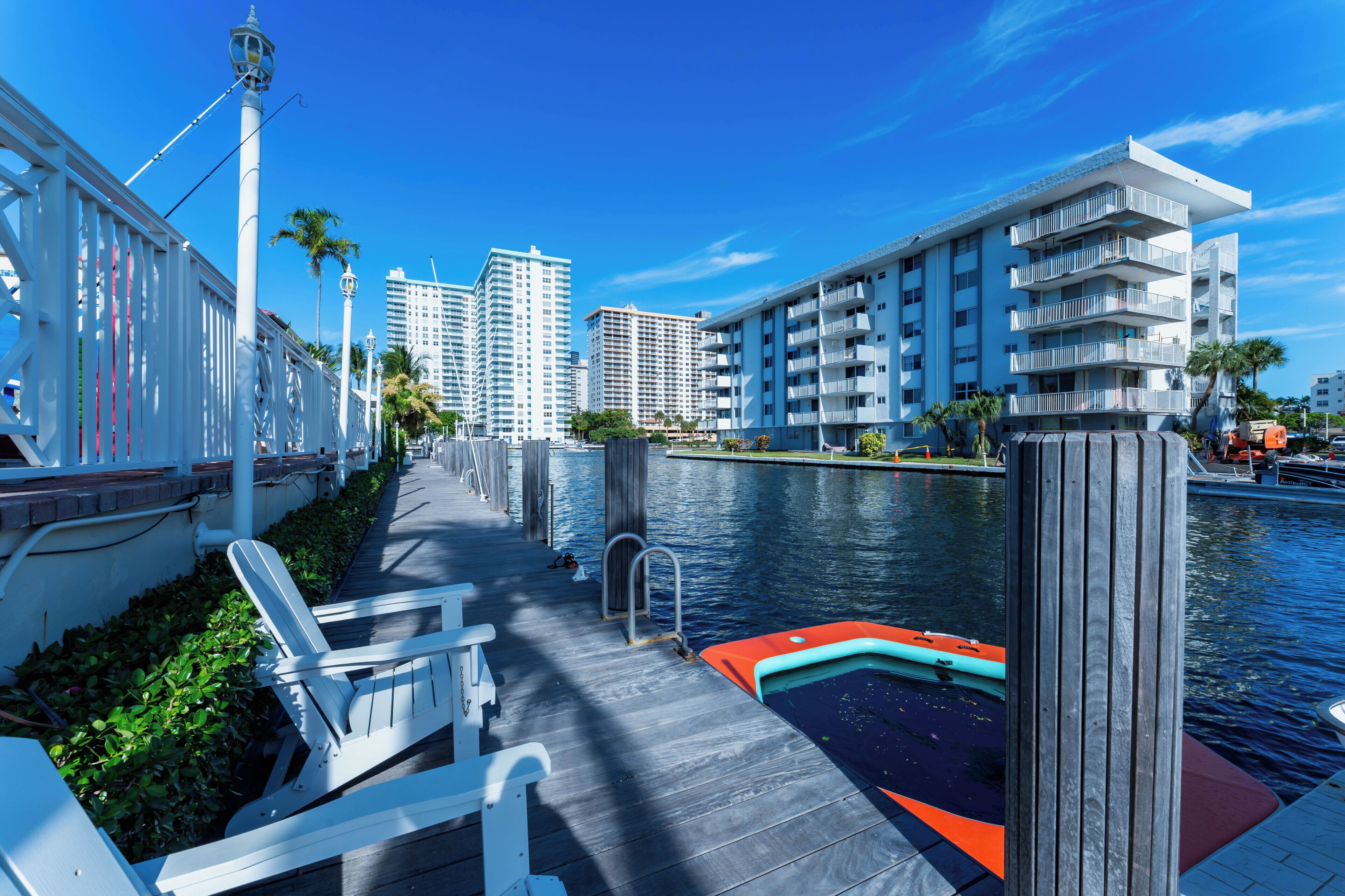 A waterfront promenade with a wooden deck, lounge chairs, and a small boat.