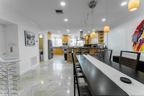 A modern kitchen with a long dining table, built-in appliances, and pendant lighting. - Luxury Waterfront W/ Heated Pool & Canal Views! (North Miami Beach)