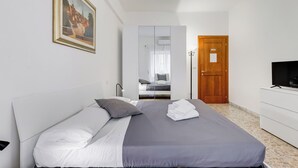 A neatly made bed with white and gray linens, a wooden door, a white dresser with a TV, and a framed picture on the wall.