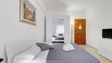 Een netjes opgemaakt bed met witte en grijze lakens, een houten deur, een witte dressoir met een televisie, en een ingelijst foto aan de muur.