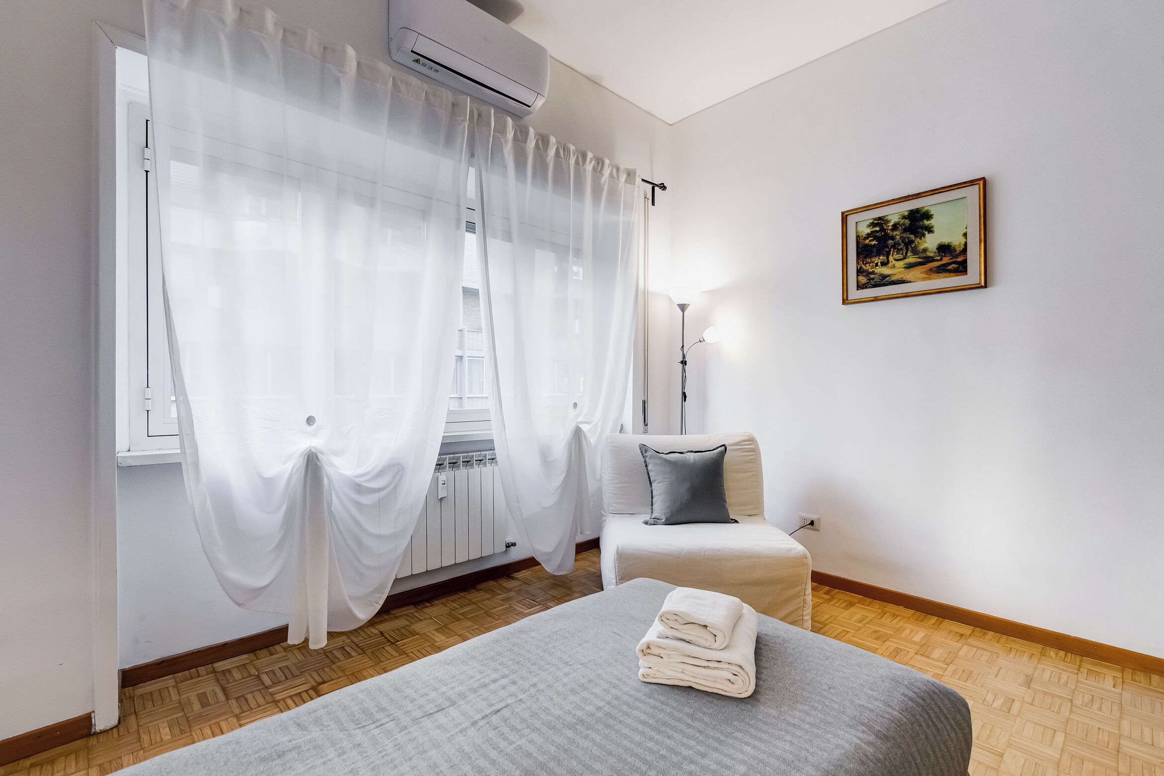 Ein Schlafzimmer mit einem Bett, einem Nachttisch, einer Lampe, einem Fenster mit Vorhängen und einem Bild an der Wand.