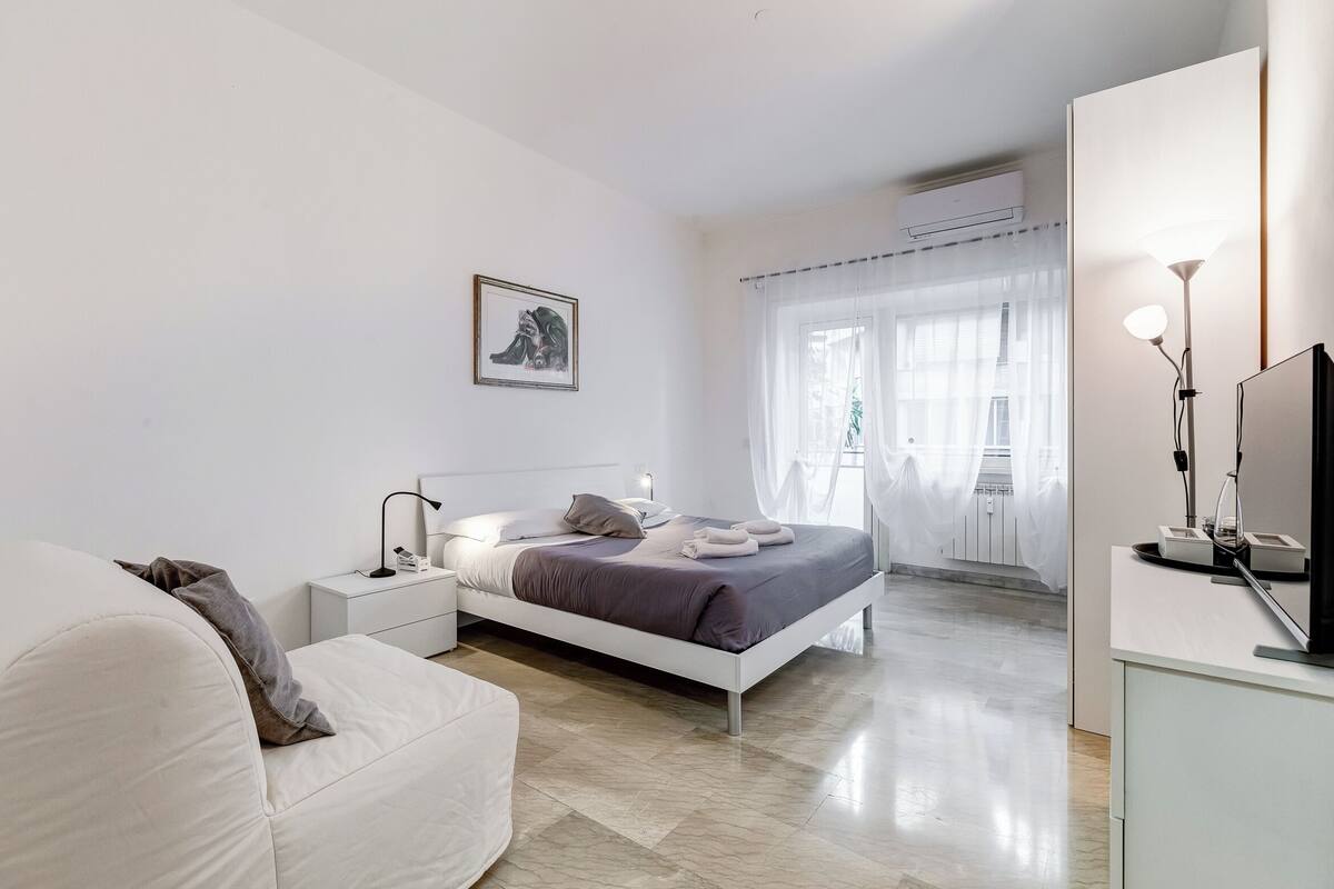 A modern bedroom with a bed, bedside tables, a TV, and a window with sheer curtains.