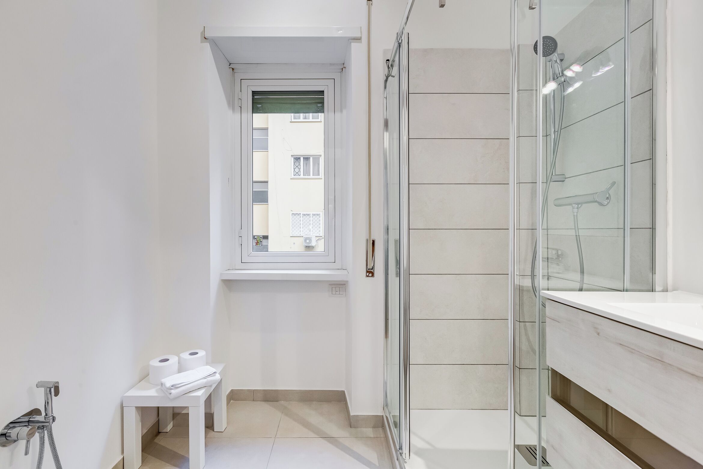 A modern bathroom with a glass shower enclosure, a small table with towels, and a window providing natural light.
