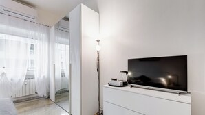 A modern hotel room with a bed, a flat-screen TV mounted on a white cabinet, a floor lamp, and a large mirror reflecting the room.
