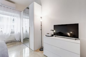 Et moderne hotelværelse med en seng, et fladskærms-TV monteret på et hvidt skab, en gulvlampe og et stort spejl, der viser rummet.