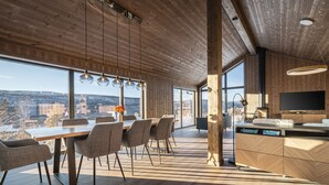 Et moderne interiør med træloft, store vinduer, spiseplads med stole, et køkken med opvaskemaskine og et fjernsyn.
